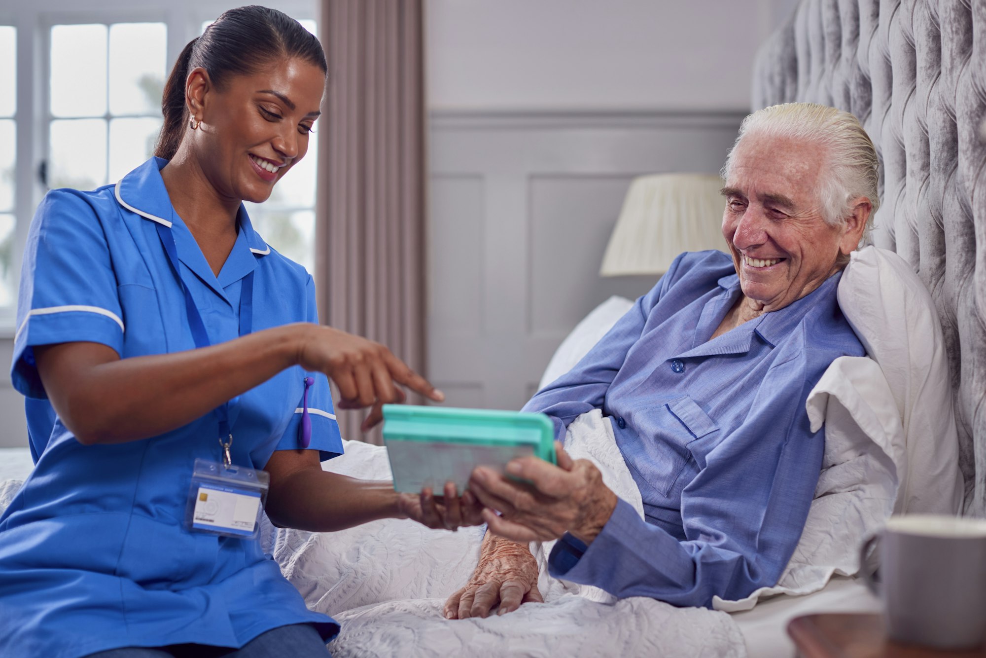 Female Care Worker In Uniform Helping Senior Man At Home In Bed With Medication