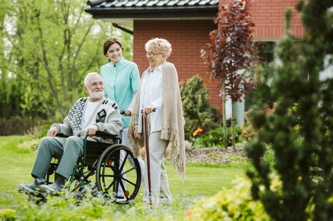 Senior guy in a wheelchair in the garden, accompanied by a home care service worker.